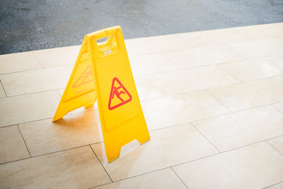 A yellow wet floor warning sign stand on a beige ceramic tiled floor in an indoor hallway or lobby area, alerting pedestrians to a potentially slippery surface. The sign features red triangle icons wi
