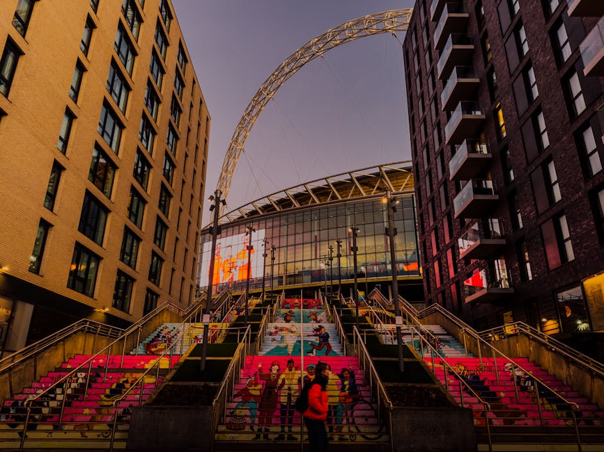 A panoramic view of Wembley Stadium's interior, showcasing the tiered red seating sections arranged in a semi-circular layout around the grass playing field. The well-maintained natural grass pitch di